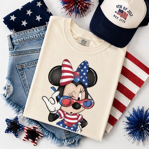 Może przedstawiać: Kremowa koszulka z grafiką Minnie Mouse w patriotycznych barwach, w tym czerwoną, białą i niebieską kokardą oraz okularami przeciwsłonecznymi. Obraz zawiera r&oacute;wnież dżinsowe szorty, granatową czapkę z napisem "4th of July" i okulary przeciwsłoneczne w kształcie gwiazd.
