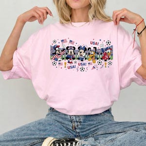 Pu&ograve; includere: T-shirt rosa chiaro con una grafica di personaggi Disney vestiti con abiti patriottici, con la scritta "USA!" e palloni da calcio. I personaggi includono Pippo, Topolino, Minnie, Pluto e Paperino.