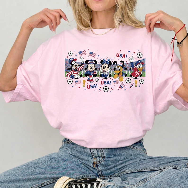 Pu&ograve; includere: T-shirt rosa chiaro con una grafica di personaggi Disney vestiti con abiti patriottici, con la scritta "USA!" e palloni da calcio. I personaggi includono Pippo, Topolino, Minnie, Pluto e Paperino.