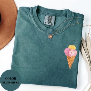 May include: A teal Comfort Colors t-shirt with an ice cream cone graphic. The ice cream cone has pink and yellow scoops. A gold necklace with a pendant is draped on the shirt. The shirt has a small tag that says "Comfort Colors".