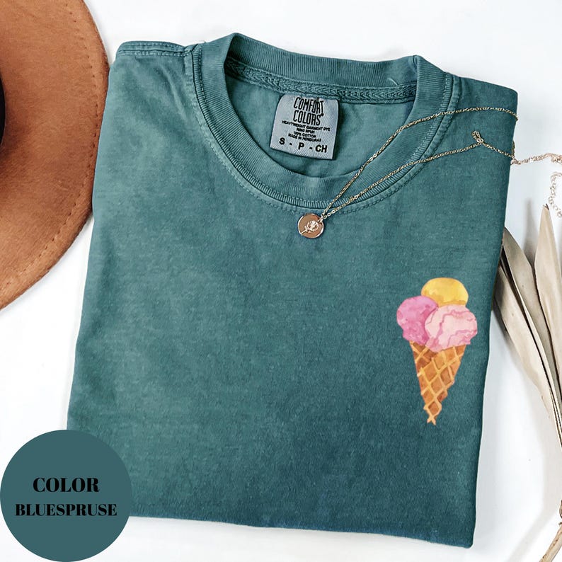 May include: A teal Comfort Colors t-shirt with an ice cream cone graphic. The ice cream cone has pink and yellow scoops. A gold necklace with a pendant is draped on the shirt. The shirt has a small tag that says "Comfort Colors".