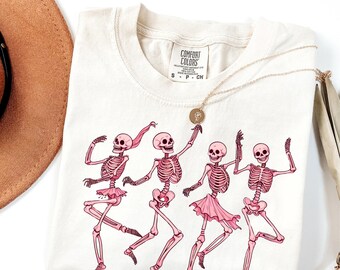 Puede incluir: Camiseta color crema con un equipo de baile de esqueletos rosas. Los esqueletos llevan tutús y cintas para el pelo rosas. La camiseta tiene la etiqueta de la marca Comfort Colors. Un collar dorado con un colgante está drapeado sobre la camiseta.