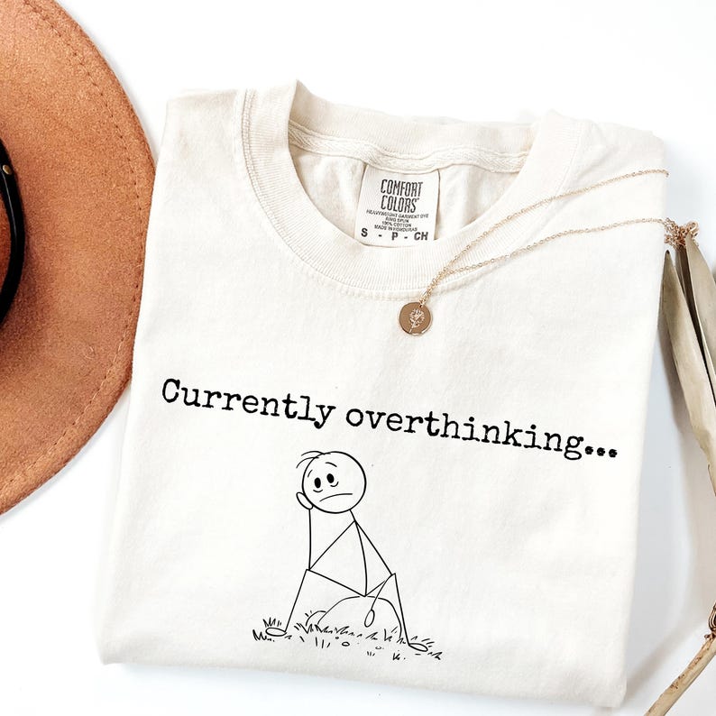 K&ouml;nnte beinhalten: Cremefarbenes T-Shirt mit dem Text "Currently overthinking..." und einer Strichm&auml;nnchen-Grafik. Das Shirt hat einen Rundhalsausschnitt und kurze &Auml;rmel. Eine goldene Halskette mit Anh&auml;nger liegt &uuml;ber dem Shirt.