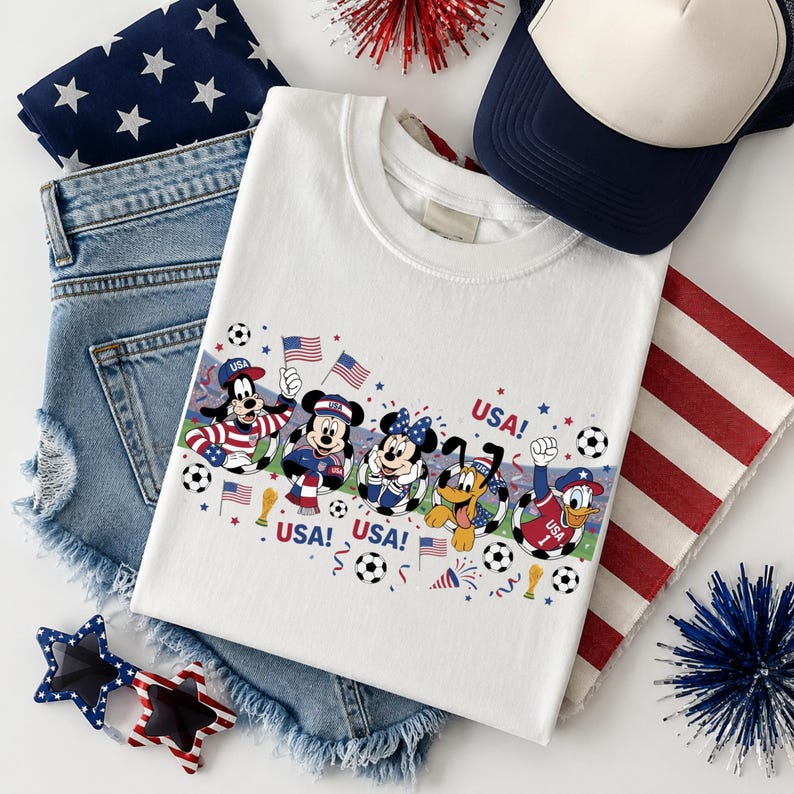 Pu&ograve; includere: Maglietta bianca con personaggi Disney in abiti patriottici, la parola "USA" e palloni da calcio. L'immagine include anche pantaloncini di jeans, un cappellino da camionista blu navy e bianco e occhiali da sole a forma di stella.