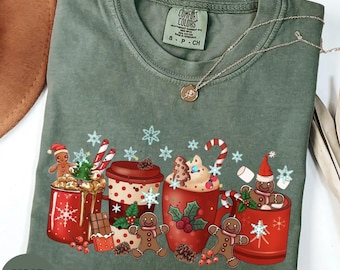 Puede incluir: Una camiseta Comfort Colors verde musgo con un diseño navideño. El gráfico incluye tazas rojas llenas de chocolate caliente, hombres de jengibre, bastones de caramelo y copos de nieve. Un collar plateado con un colgante está sobre la camiseta.