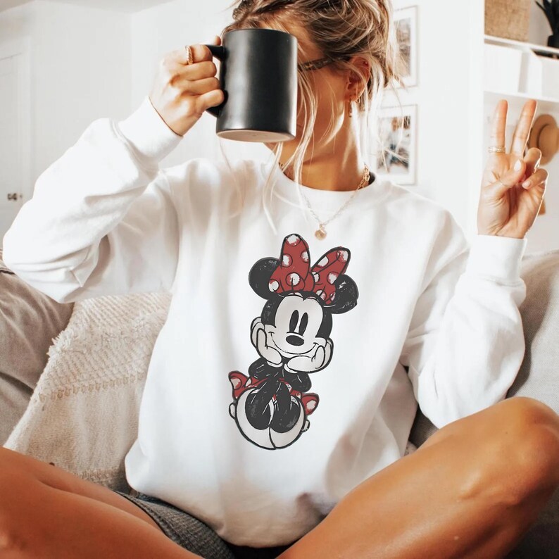 May include: White sweatshirt featuring a Minnie Mouse graphic with a red polka dot bow. The cartoon character is in black and white. The sweatshirt is a casual style, suitable for everyday wear.