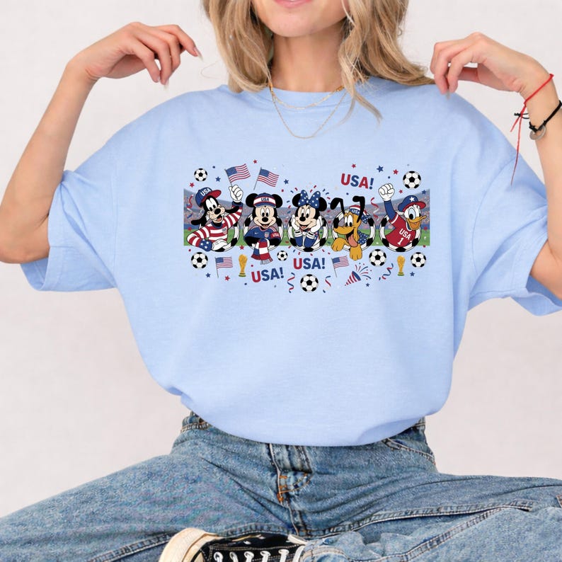Pu&ograve; includere: T-shirt azzurra con una grafica di personaggi Disney in tenuta da calcio, che sventolano bandiere americane. Il testo "USA!" &egrave; visualizzato sopra e sotto i personaggi. Sono inclusi anche palloni da calcio e coriandoli.