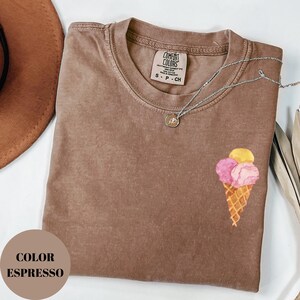 May include: Espresso-colored t-shirt with a small ice cream cone graphic. The cone has pink and yellow scoops. A silver necklace with a small pendant rests on the shirt. The shirt has a Comfort Colors label.