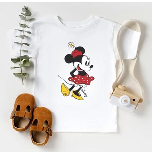 Puede incluir: Camiseta blanca con un dibujo de Minnie Mouse con falda de lunares rojos, lazo rojo y zapatos amarillos. La palabra "Disney" est&aacute; impresa debajo. Zapatos de beb&eacute; de cuero marr&oacute;n y una correa crema tambi&eacute;n est&aacute;n en la imagen.