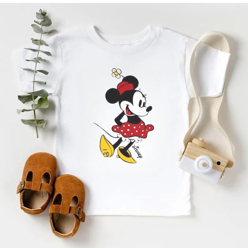 Puede incluir: Camiseta blanca con un dibujo de Minnie Mouse con falda de lunares rojos, lazo rojo y zapatos amarillos. La palabra "Disney" est&aacute; impresa debajo. Zapatos de beb&eacute; de cuero marr&oacute;n y una correa crema tambi&eacute;n est&aacute;n en la imagen.