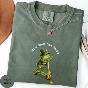 May include: A moss green t-shirt with the phrase "Off to Commit Irish Foolery" encircling a cartoon frog riding a scooter. The frog wears a green hat and jacket. A gold-colored necklace with a pendant is draped across the shirt.