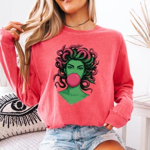 May include: Coral-colored sweatshirt featuring a graphic of Medusa with green skin, pink hair, and a bubblegum bubble. The illustration has a pop art style. The sweatshirt has long sleeves and a crew neck.