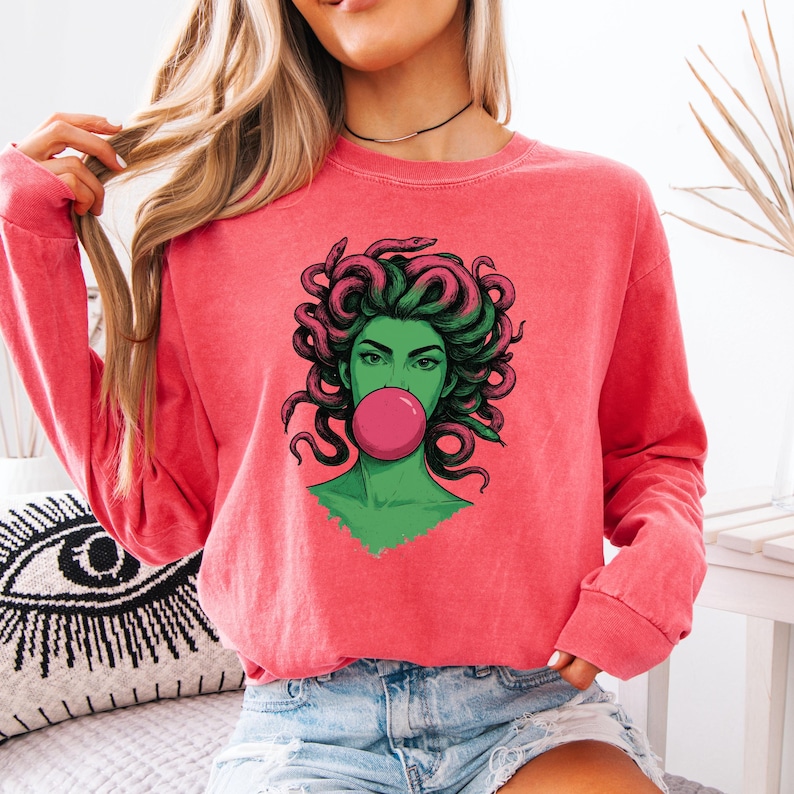 May include: Coral-colored sweatshirt featuring a graphic of Medusa with green skin, pink hair, and a bubblegum bubble. The illustration has a pop art style. The sweatshirt has long sleeves and a crew neck.