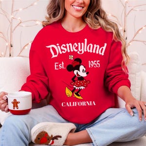 May include: A red Disneyland sweatshirt with a Minnie Mouse graphic in a polka dot dress. The sweatshirt displays "Disneyland Est. 1955 CALIFORNIA" in white. A white mug with a gingerbread man is held in the hand.