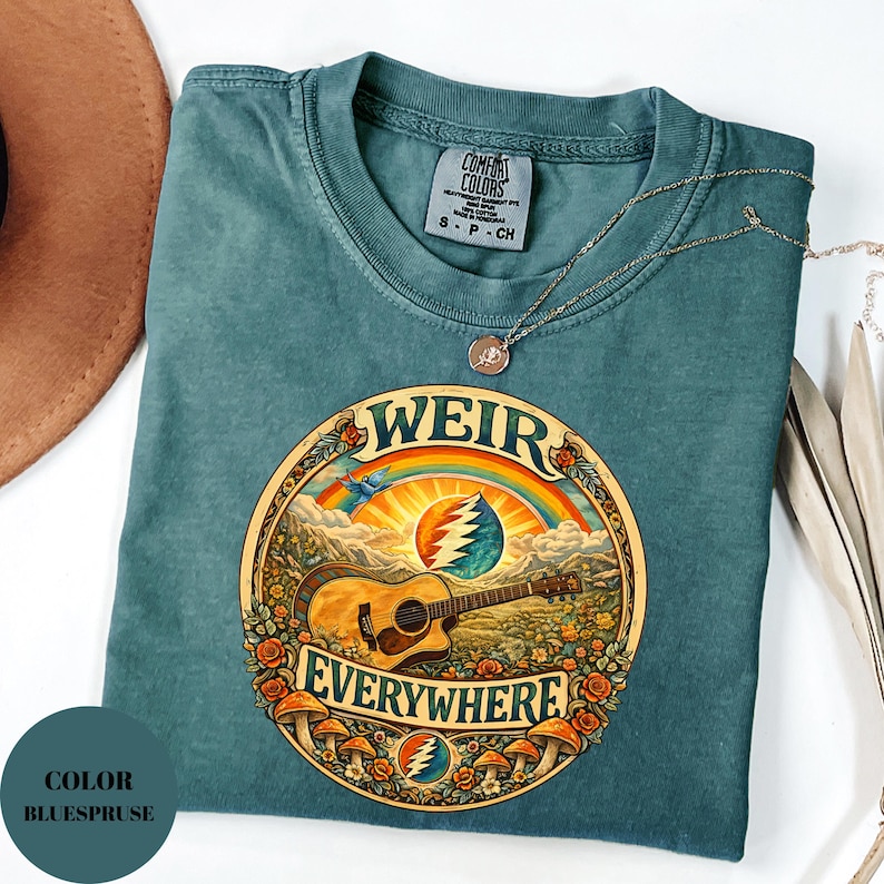 May include: A teal t-shirt with a circular graphic featuring a guitar, rainbow, and the words "WEIR EVERYWHERE." The shirt has a crew neck and short sleeves. A gold necklace is draped across the shirt.