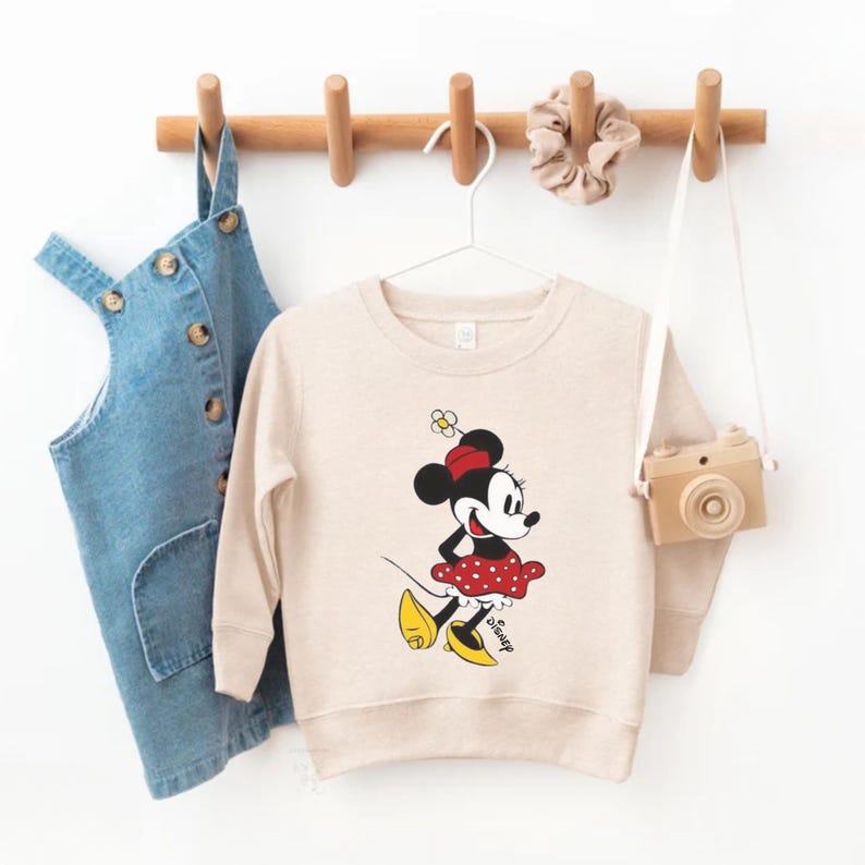 Puede incluir: Sudadera beige con un dise&ntilde;o de Minnie Mouse. Minnie aparece con un vestido de lunares rojos, un lazo rojo y zapatos amarillos. La palabra "Disney" est&aacute; impresa debajo. Un peto vaquero y una c&aacute;mara de juguete cuelgan cerca.