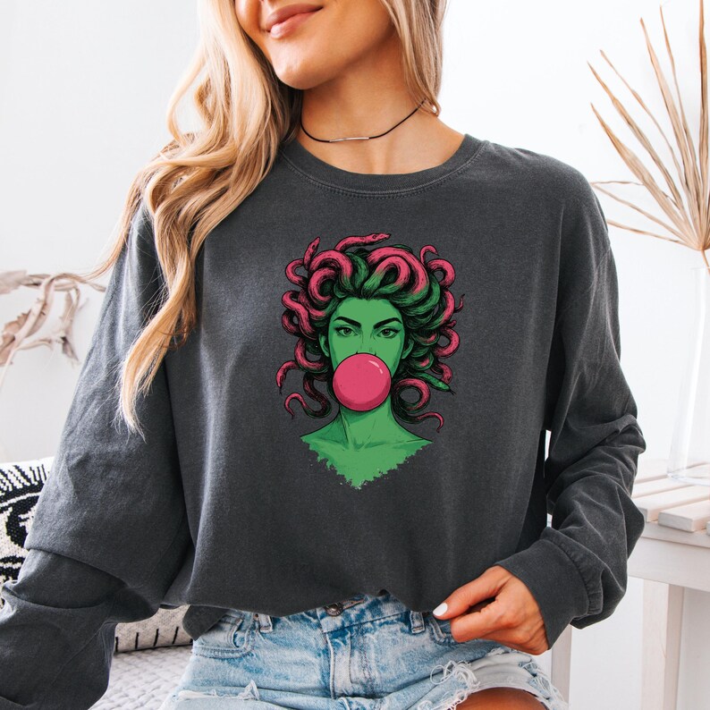 May include: Dark gray long-sleeve sweatshirt featuring a graphic of Medusa with green skin, pink hair, and a pink bubblegum bubble. The graphic is centered on the front of the sweatshirt. The model is wearing denim shorts.