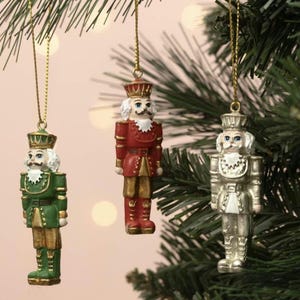 May include: Three hanging nutcracker ornaments featuring detailed uniforms and hats. One is painted green and gold, one is red and gold, and one is metallic silver. Each figure has a white beard and moustache and hangs from a gold string.