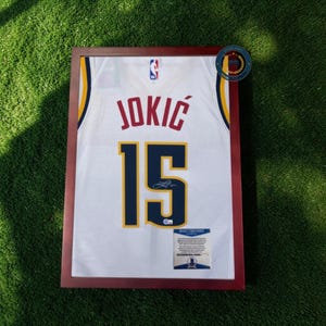 Może przedstawiać: Oprawiona biała koszulka do koszykówki z napisem "JOKIC" i numerem "15" w kolorze granatowym. Koszulka ma żółte i granatowe akcenty oraz logo NBA. Rama jest w ciemnobrązowym kolorze. Widoczny jest certyfikat autentyczności.