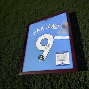 Pu&ograve; includere: Maglia da calcio azzurra incorniciata con il nome "HAALAND" e il numero "9" in bianco. La maglia &egrave; firmata e include un "BECKETT CERTIFICATE" di autenticit&agrave;. La cornice &egrave; marrone scuro.