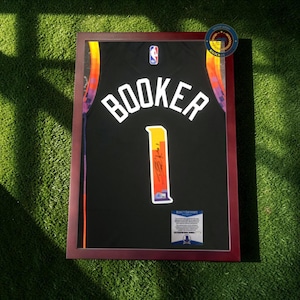 May include: Framed black basketball jersey with the name "BOOKER" and the number "1" in white. The jersey has a colorful design on the sides and is set in a dark red frame. A certificate is visible at the bottom.