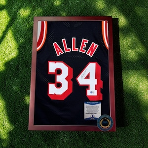 Może przedstawiać: Oprawiona czarna koszulka z nazwiskiem "ALLEN" i numerem "34" w kolorze czerwono-białym. Koszulka jest prezentowana w brązowej drewnianej ramie. Widoczny jest certyfikat autentyczności. Koszulka leży na zielonej powierzchni.