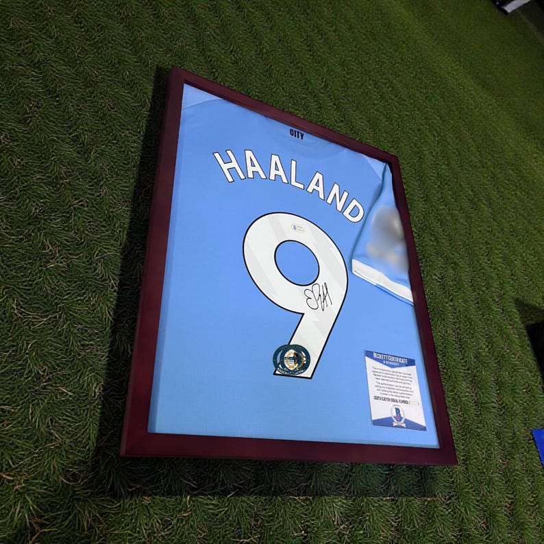 Pu&ograve; includere: Maglia da calcio azzurra incorniciata con il nome "HAALAND" e il numero "9" in bianco. La maglia &egrave; firmata e include un certificato di autenticit&agrave;. La cornice &egrave; marrone scuro, su sfondo verde.