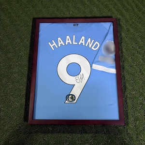 Pu&ograve; includere: Maglia da calcio azzurra incorniciata con il nome "HAALAND" e il numero "9" in bianco. La maglia &egrave; firmata e presentata in una cornice marrone scuro. La parola "CITY" &egrave; in alto.