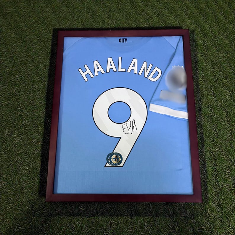 Pu&ograve; includere: Maglia da calcio azzurra incorniciata con il nome "HAALAND" e il numero "9" in bianco. La maglia &egrave; firmata e presentata in una cornice marrone scuro. La parola "CITY" &egrave; in alto.