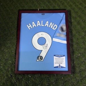 Pu&ograve; includere: Maglia azzurra incorniciata con il nome "HAALAND" e il numero "9" in bianco. La maglia &egrave; firmata e include un'etichetta "BECKETT CERTIFICATE". La cornice &egrave; di colore marrone scuro.