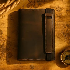 Può includere: Un diario in pelle marrone scuro con un bordo cucito e un cinturino in pelle abbinato con un elastico. Il diario è su una mappa vintage con una bussola nell'angolo.