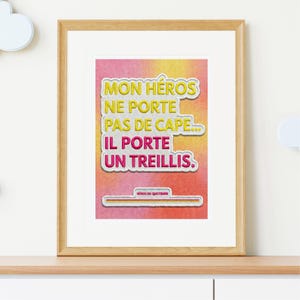 Peut inclure: Un cadre en bois contenant une impression avec le texte "MON HEROS NE PORTE PAS DE CAPE... IL PORTE UN TREILLIS." en jaune et rose. Le fond est un dégradé de rose, jaune et orange.