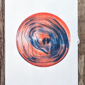 May include: Abstract art print featuring a circular design with concentric lines. The artwork uses shades of red and blue, creating a visually engaging pattern. The print is on white paper, set against a rustic wooden background.