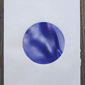 May include: A white paper with a large, detailed blue circle in the center. The circle has a textured, swirling pattern, creating a sense of depth and movement. The paper is set against a wooden background.