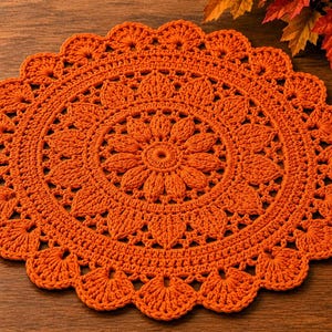 May include: A round, bright orange crochet doily featuring a detailed, repeating floral-inspired pattern with a central starburst design and a scalloped, shell-like edge. The intricate needlework creates a textured, circular textile piece.