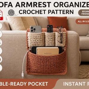 May include: A crochet sofa armrest organizer pattern in PDF format. The organizer is rust-colored with three beige upper pockets and one large lower pocket. It holds a smartphone, remote, glasses, and a notebook with a white charging cable.