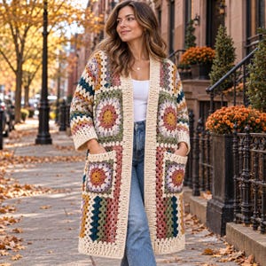 May include: A long, open-front cardigan made of multicolored crochet granny squares in shades of mustard yellow, sage green, dusty rose, and deep teal, bordered by cream-colored yarn. The garment features two large front pockets and long sleeves.