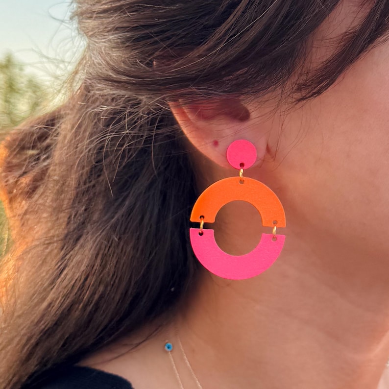 May include: Vibrant, geometric earrings in pink and orange. Each earring has a small pink circle at the top, connected to a larger, open semi-circle design. The top half is orange, and the bottom half is pink.