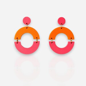 May include: A pair of colourful, geometric earrings. Each earring has a small pink circle at the top, connected to a larger orange semi-circle, and a pink semi-circle below. Gold-coloured hardware connects the pieces. The earrings are displayed on a white background.