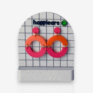 May include: Bright, geometric earrings on a white and grey grid display. Each earring has a pink top circle, an orange semi-circle, and a pink bottom semi-circle. The brand name "happiears" is displayed in blue. A green smiley face logo is also visible.