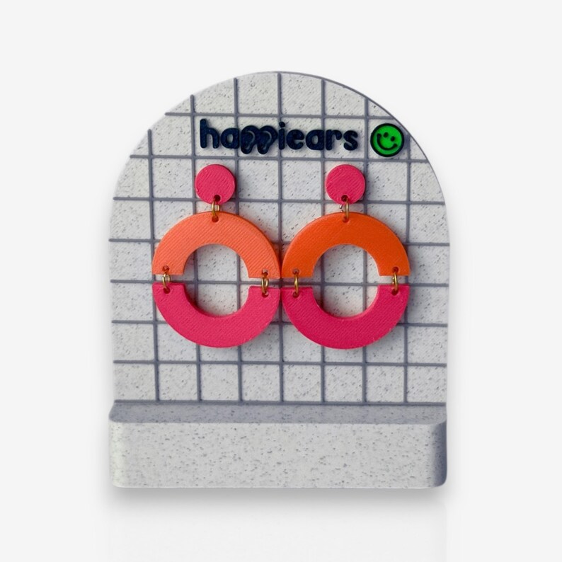 May include: Bright, geometric earrings on a white and grey grid display. Each earring has a pink top circle, an orange semi-circle, and a pink bottom semi-circle. The brand name "happiears" is displayed in blue. A green smiley face logo is also visible.