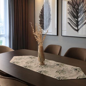 May include: A dark brown wooden dining table with a floral table runner and a vase with dried flowers. Two framed black and white botanical prints hang on the wall behind the table. Four brown upholstered chairs surround the table.
