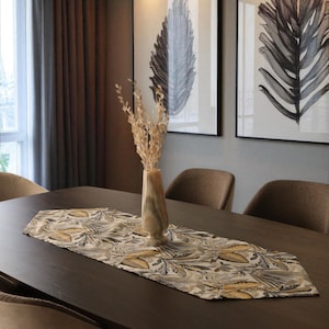 Puede incluir: Una mesa de comedor con un camino de mesa estampado, un jarrón con flores secas y cuatro sillas tapizadas. Dos estampados botánicos enmarcados cuelgan de la pared. El camino de mesa presenta un diseño de hojas en tonos amarillos, grises y beige.