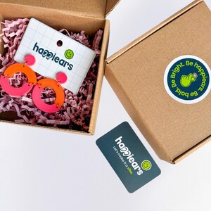 May include: A pair of pink and orange hoop earrings in a brown box with pink shredded paper. The box contains a card with the text "happiears" and a smiley face logo. A sticker on the box reads "Be Bright. Be bold. happiears."
