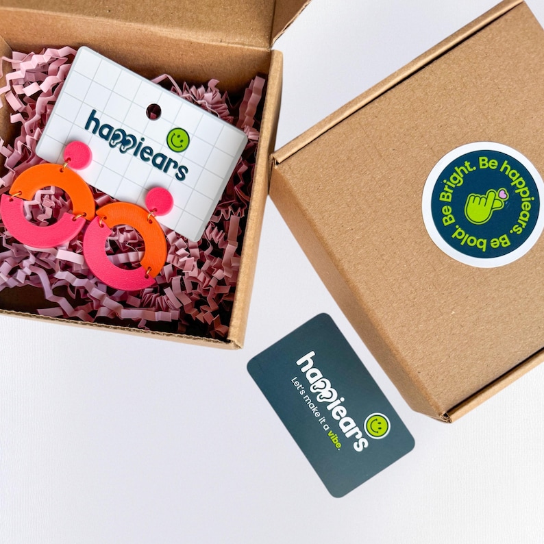 May include: A pair of pink and orange hoop earrings in a brown box with pink shredded paper. The box contains a card with the text "happiears" and a smiley face logo. A sticker on the box reads "Be Bright. Be bold. happiears."