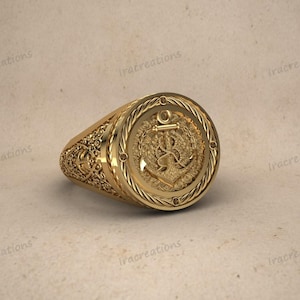 以下が含まれることがあります： 金色のリングで、錨のデザインが施されています。リングには、錨の周りのロープのような縁取りや、側面のフローラルパターンなど、複雑なディテールが施されています。リングはニュートラルな背景に表示されています。