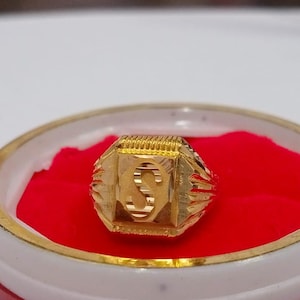 May include: A gold ring featuring the letter 'S' on a red velvet base, presented in a white and gold-edged container. The ring's face is rectangular with a textured pattern.