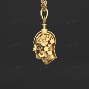 Puede incluir: Un colgante dorado con forma de cabeza humana, con engranajes y componentes mecánicos visibles en su interior. Cuelga de una cadena dorada. El diseño sugiere una temática steampunk o mecánica.