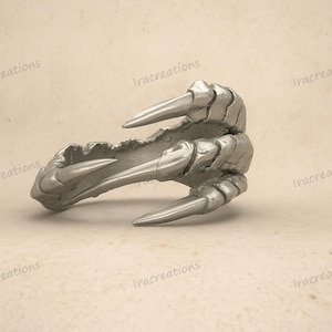 Può includere: Un anello color argento a forma di artiglio. L'anello presenta tre artigli affilati e appuntiti che si estendono dal corpo principale. Il design presenta dettagli strutturati, conferendogli un aspetto realistico. L'anello è posto su uno sfondo neutro.