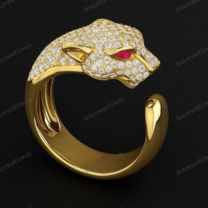 May include: A gold ring with a panther head design. The panther's head is covered in small, sparkling white stones, with a ruby-coloured stone for the eye. The ring has a smooth, polished band.